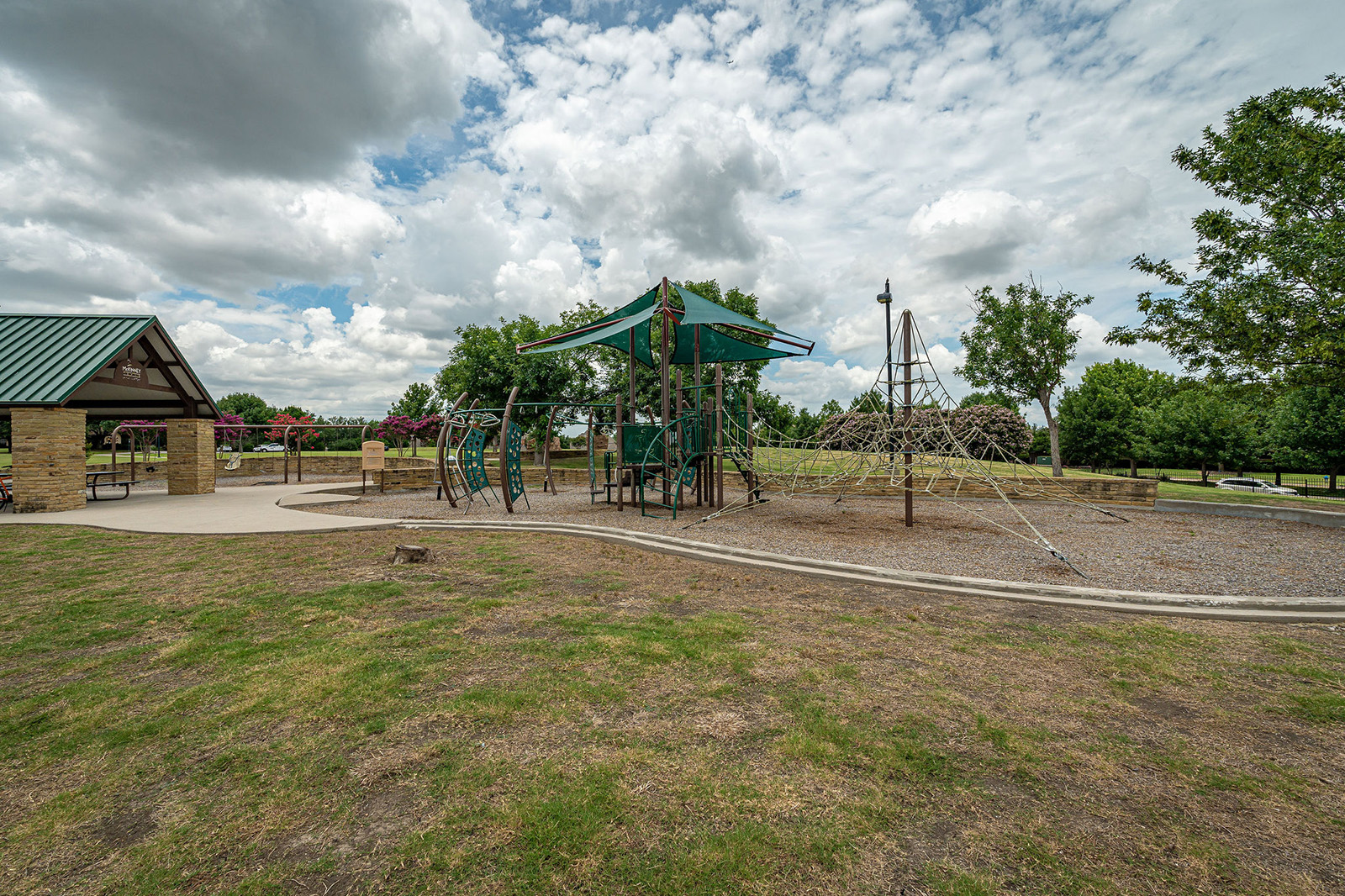 A playground with climbing structures and slides sits next to a covered pavilion in a park with green grass, trees, and a partly cloudy sky.