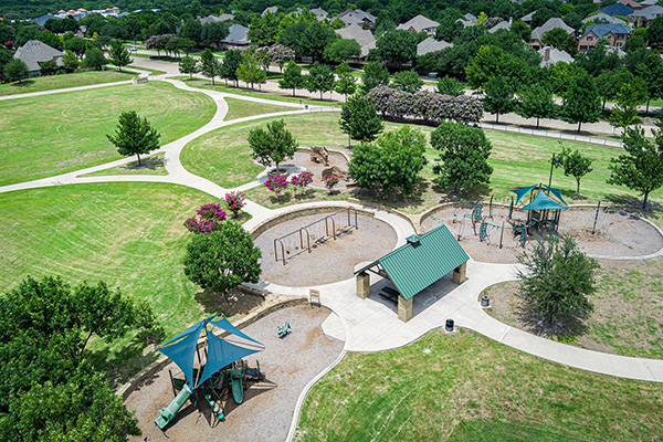 Aerial view of a suburban park with walking paths, playground equipment, a covered pavilion, trees, and surrounding residential houses.