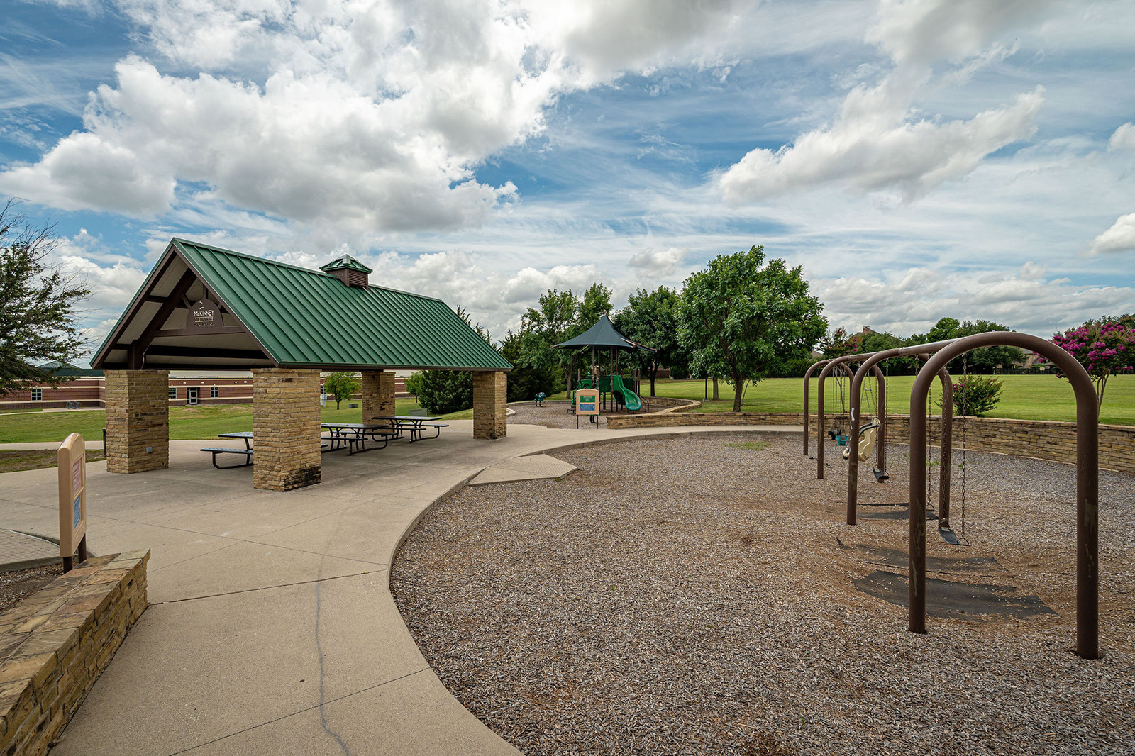 A park scene showing a covered picnic area with tables, a playground with slides, and a swing set, surrounded by grass and trees under a partly cloudy sky.