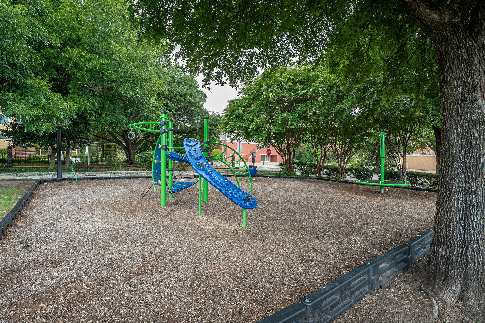 Outdoor playground area with blue and green exercise equipment surrounded by trees and woodchip ground covering.