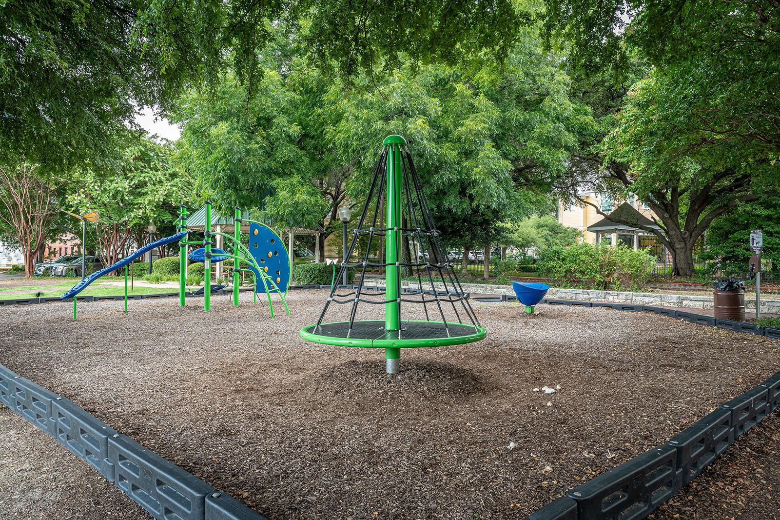 Playground with green climbing equipment, a rotating net structure, slide, spinner, and wood chip ground, surrounded by trees and a border.