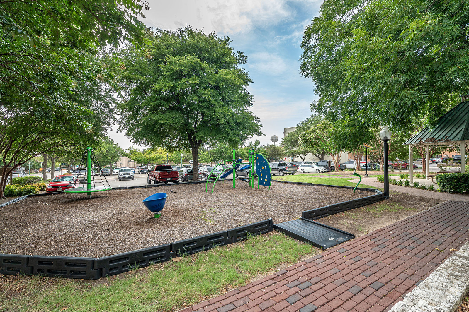 Small playground with climbing structures and swings surrounded by trees, located next to a parking lot and brick walkway.
