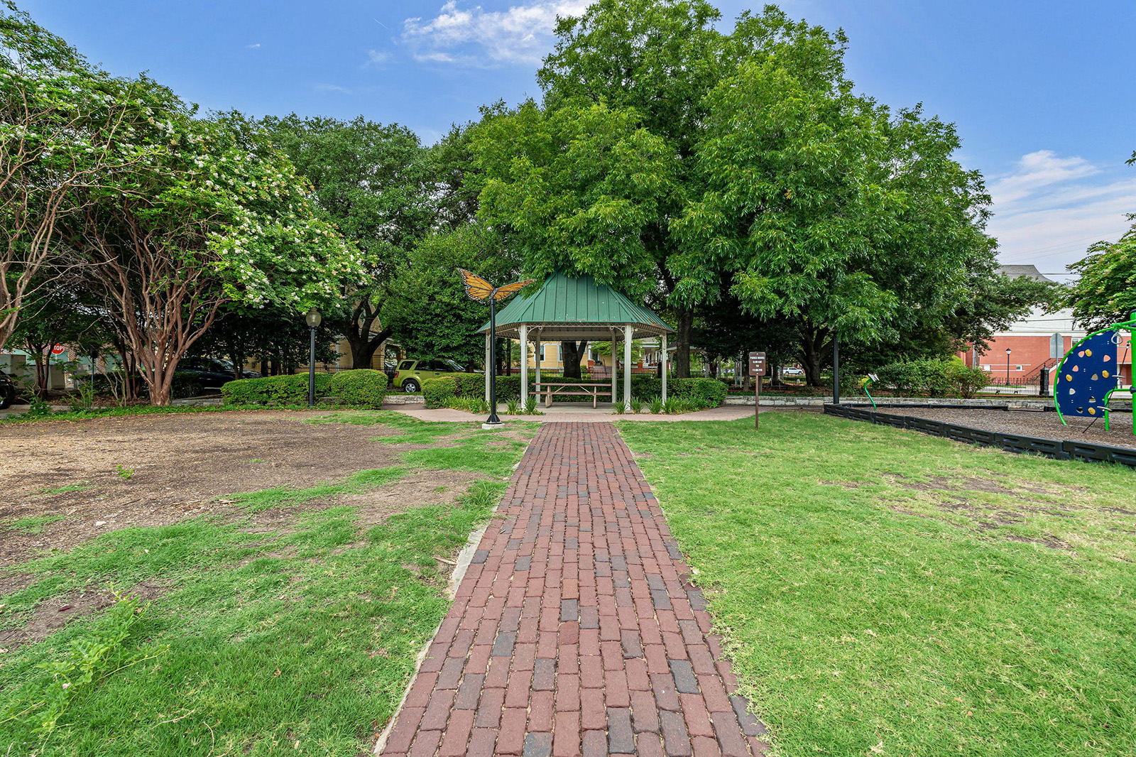 A brick pathway leads to a green-roofed gazebo surrounded by trees in a park, with a playground visible on the right.