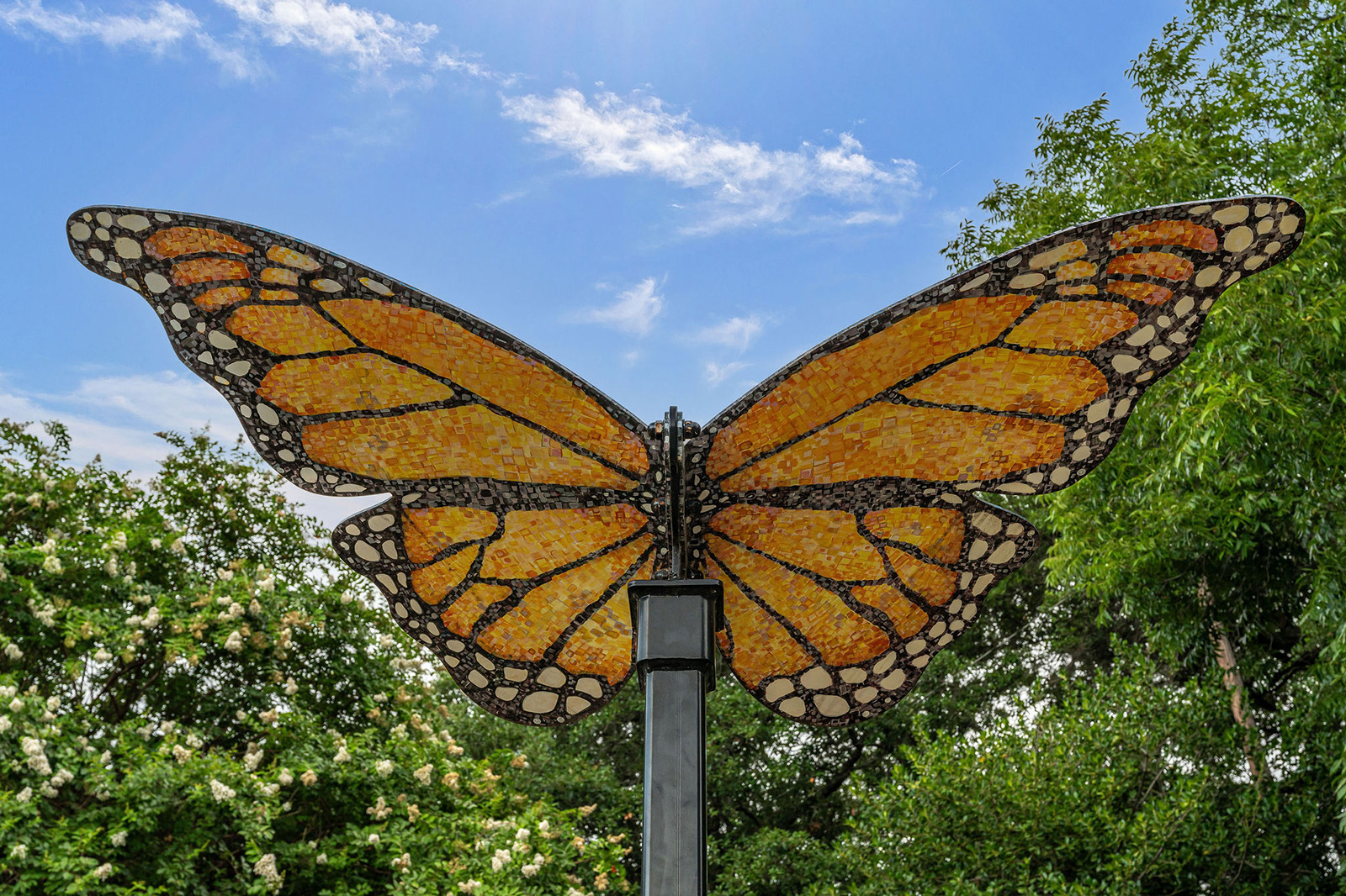 A large metal sculpture of a monarch butterfly's wings on a pole, with trees and a blue sky in the background.