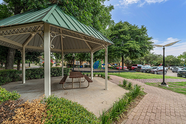 A metal gazebo with a picnic table underneath sits in a park near a parking lot, surrounded by trees, hedges, and a brick walkway.