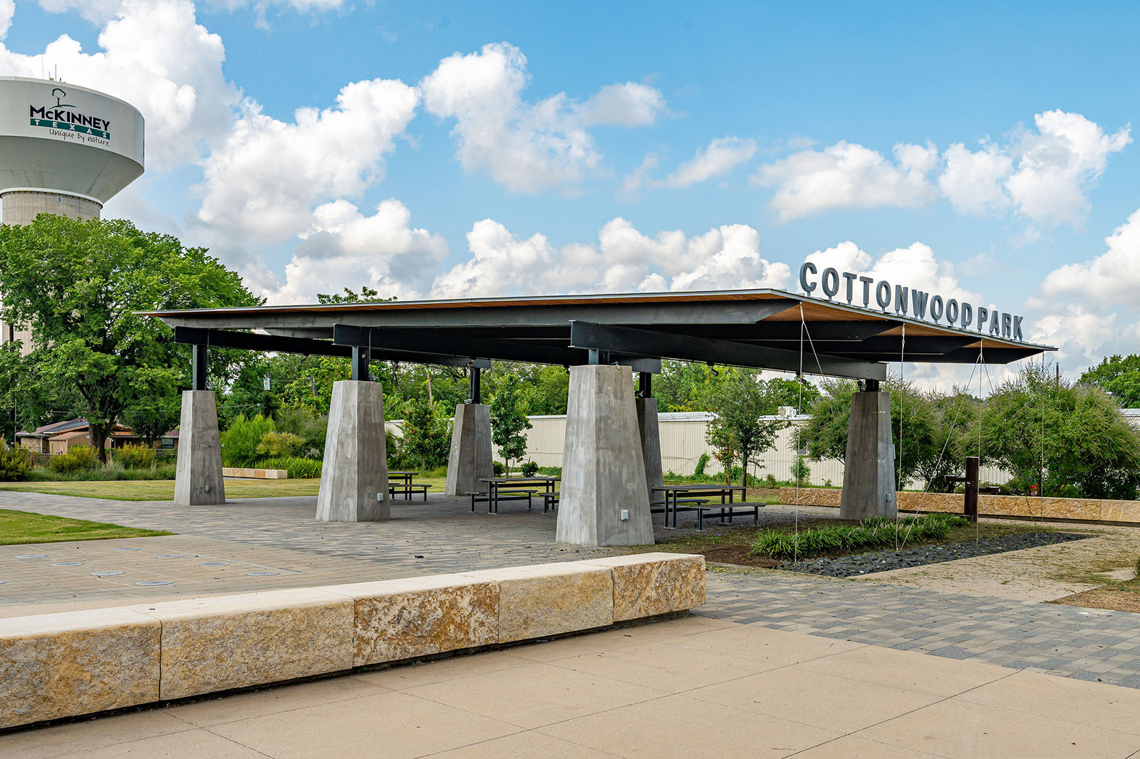 A modern pavilion with picnic tables stands beneath a sign reading "Cottonwood Park," with a McKinney water tower visible in the background.
