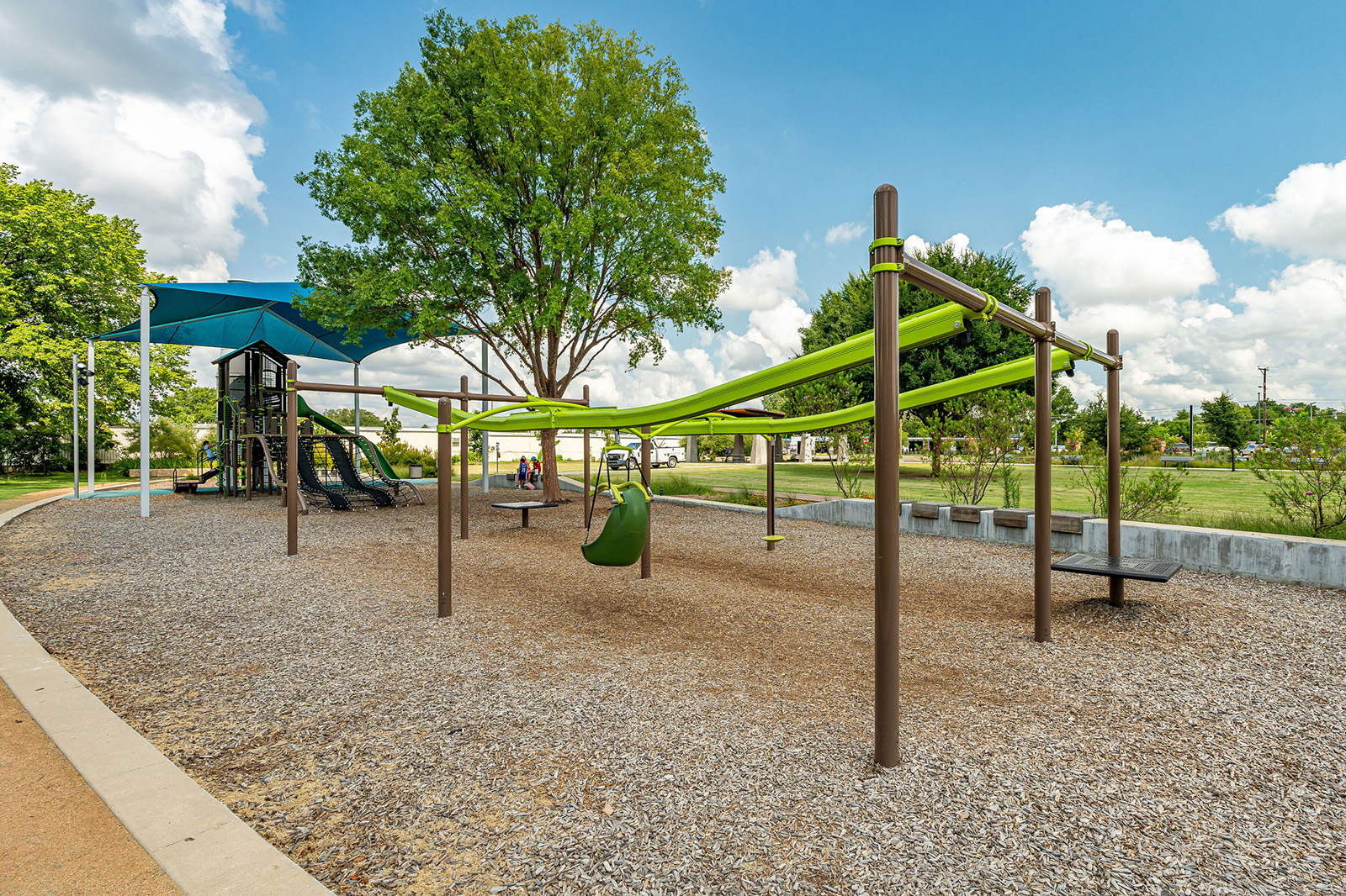 A playground with a green zip line, slide, climbing structure, trees, and benches under a blue sky with scattered clouds.