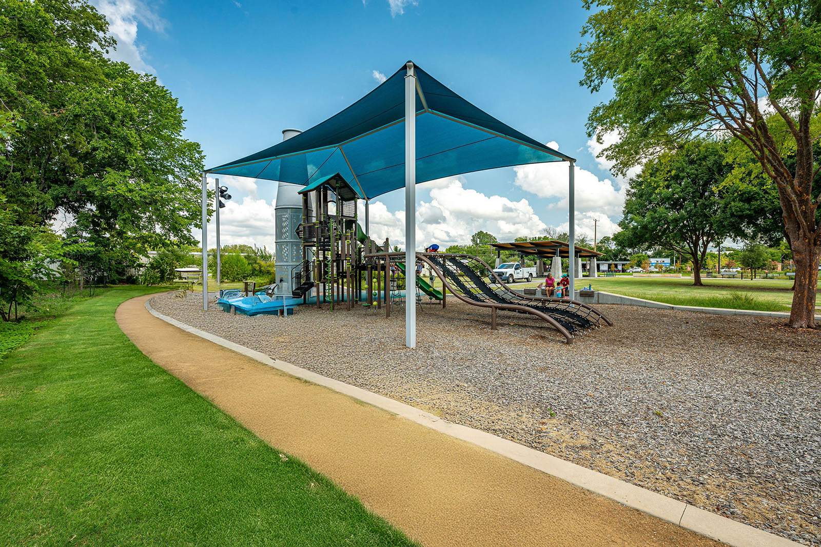 A playground with slides and climbing structures under a large blue canopy, next to a walking path and surrounded by green grass and trees.