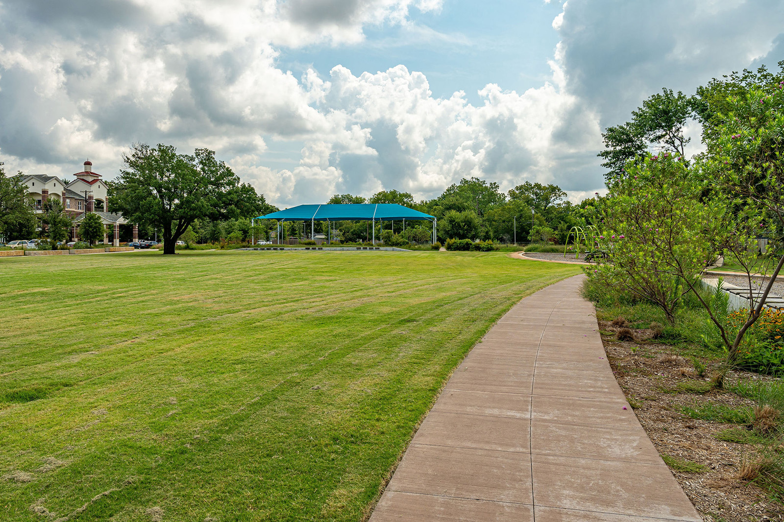 A concrete path curves through a grassy park with scattered trees, a blue-roofed pavilion, and partly cloudy skies overhead.