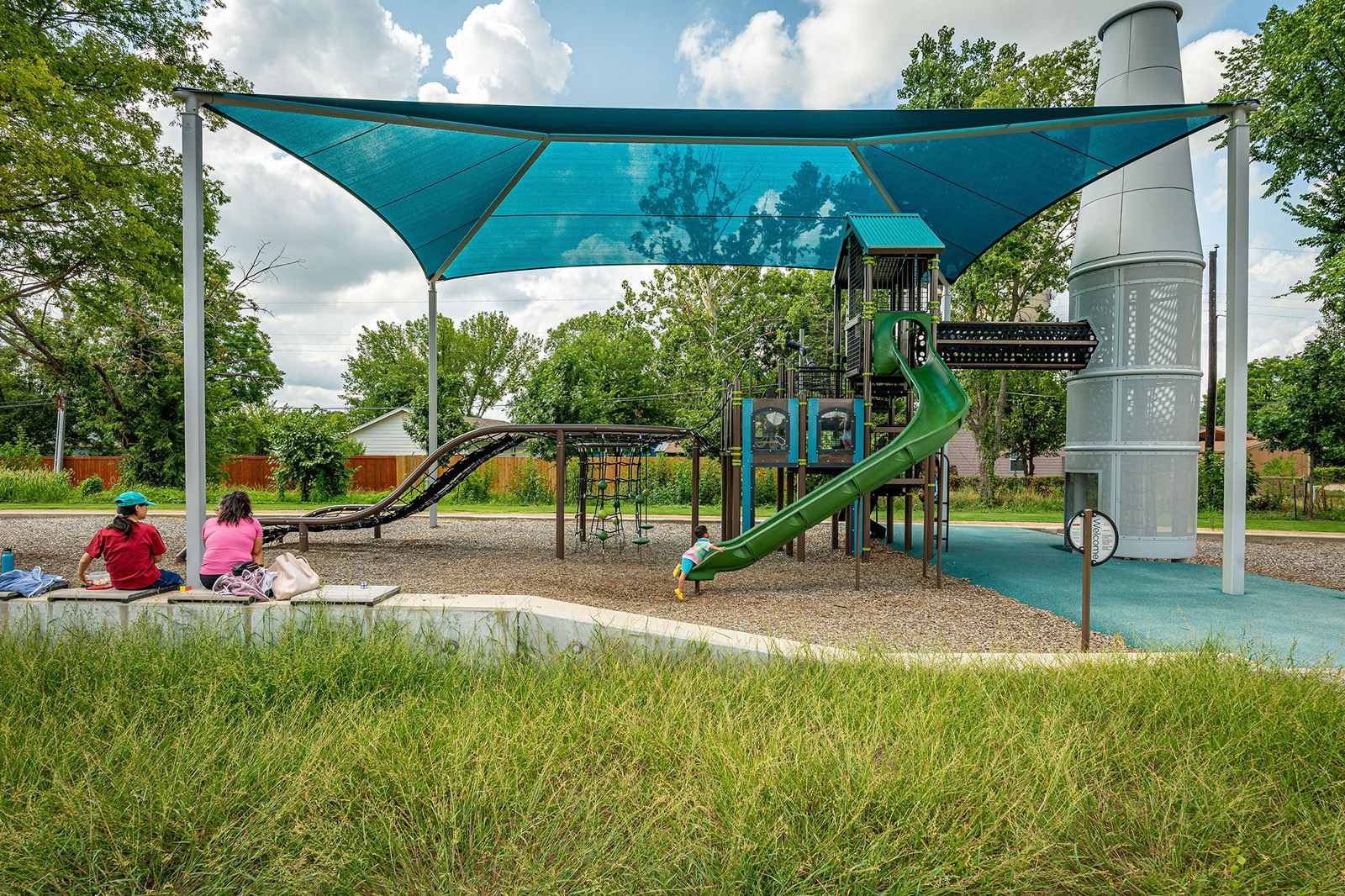 Children sit on a bench beside a modern playground with climbing structures, a green slide, and a blue sun shade, surrounded by trees and grass on a partly cloudy day.