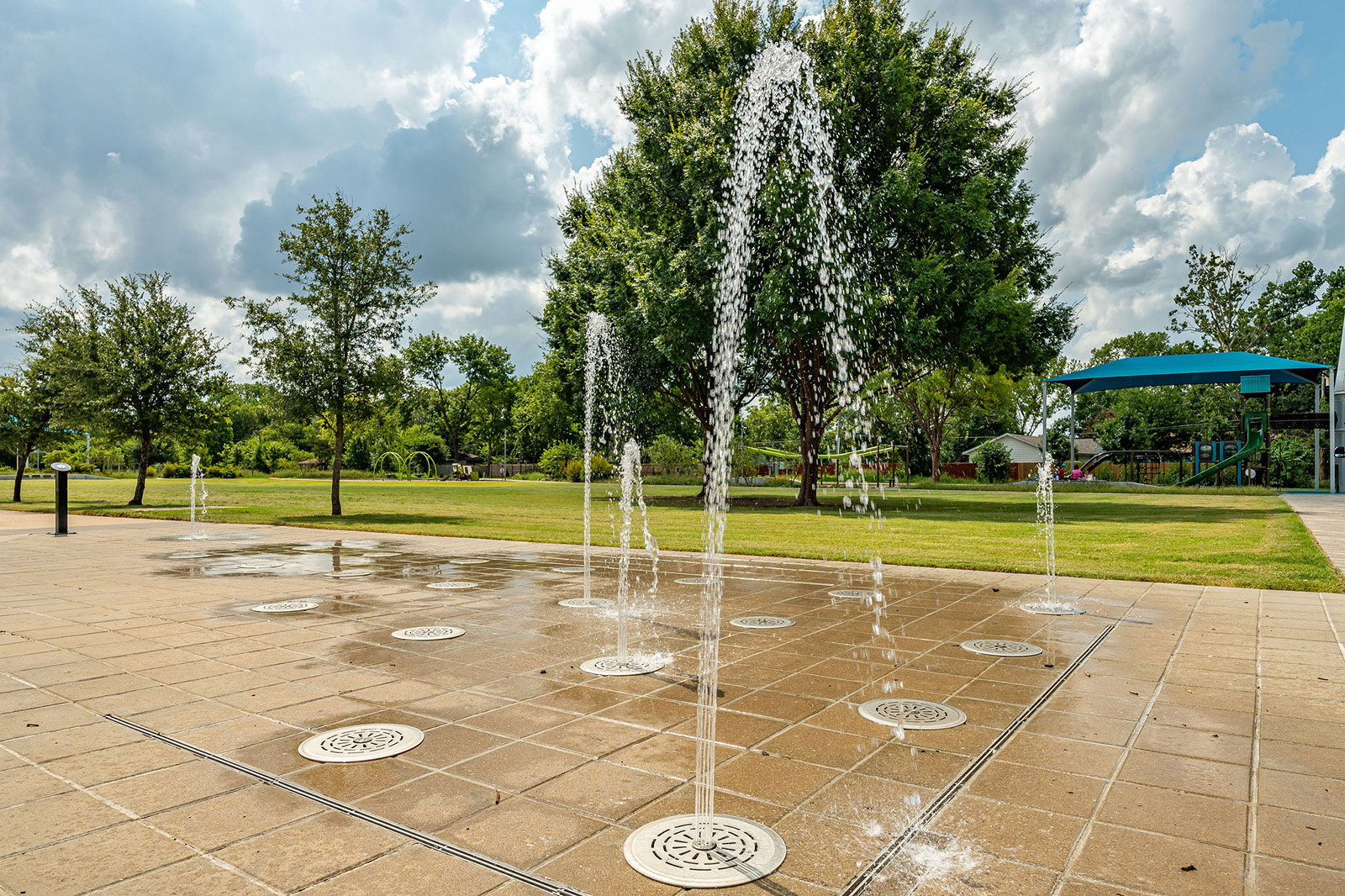 Water jets spray upward from ground-level fountains in a tiled splash pad area at a park, with trees and a blue canopy in the background.