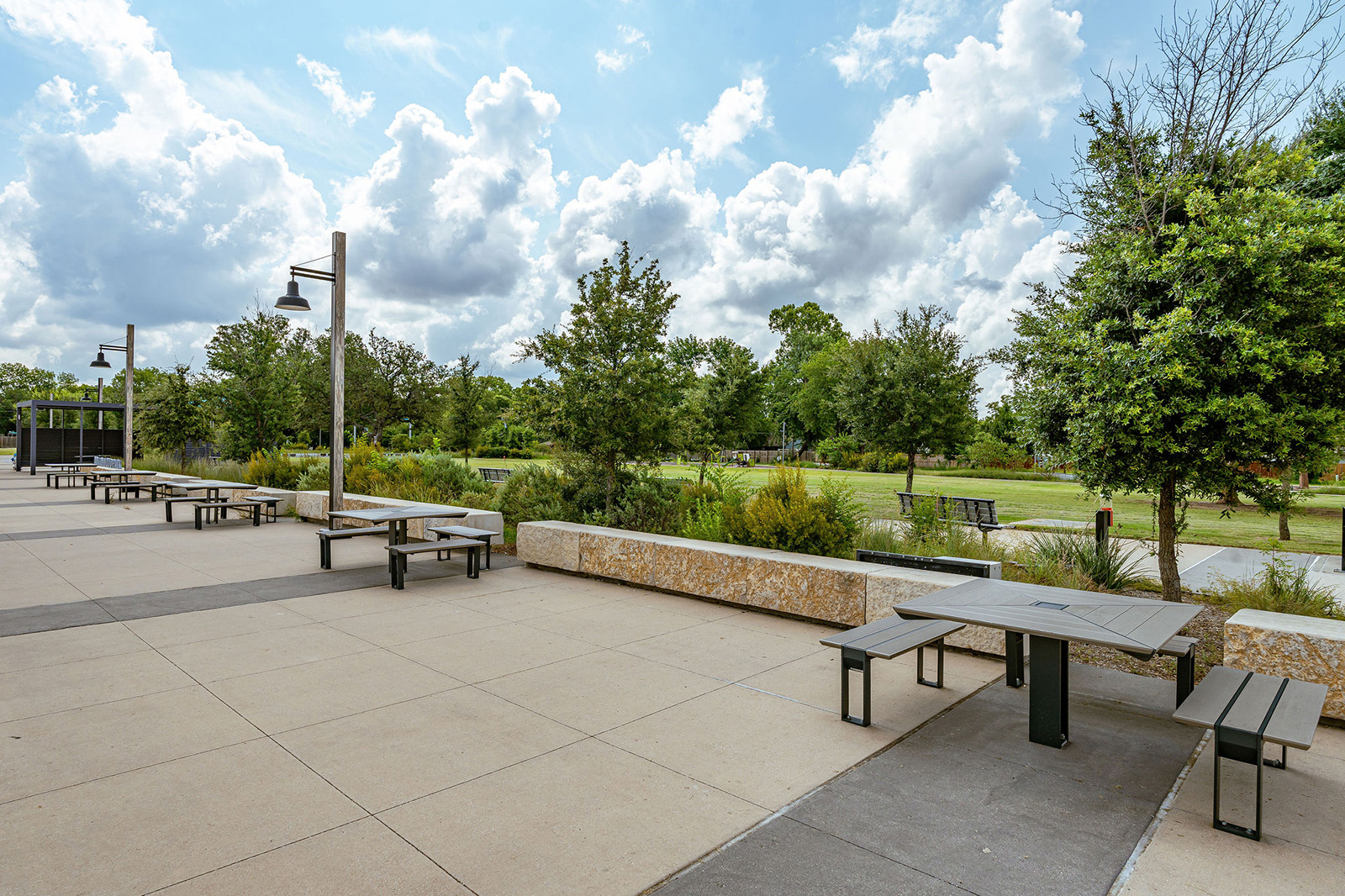 Outdoor patio area with multiple picnic tables and benches, bordered by low stone walls and landscaped greenery, under a partly cloudy sky.