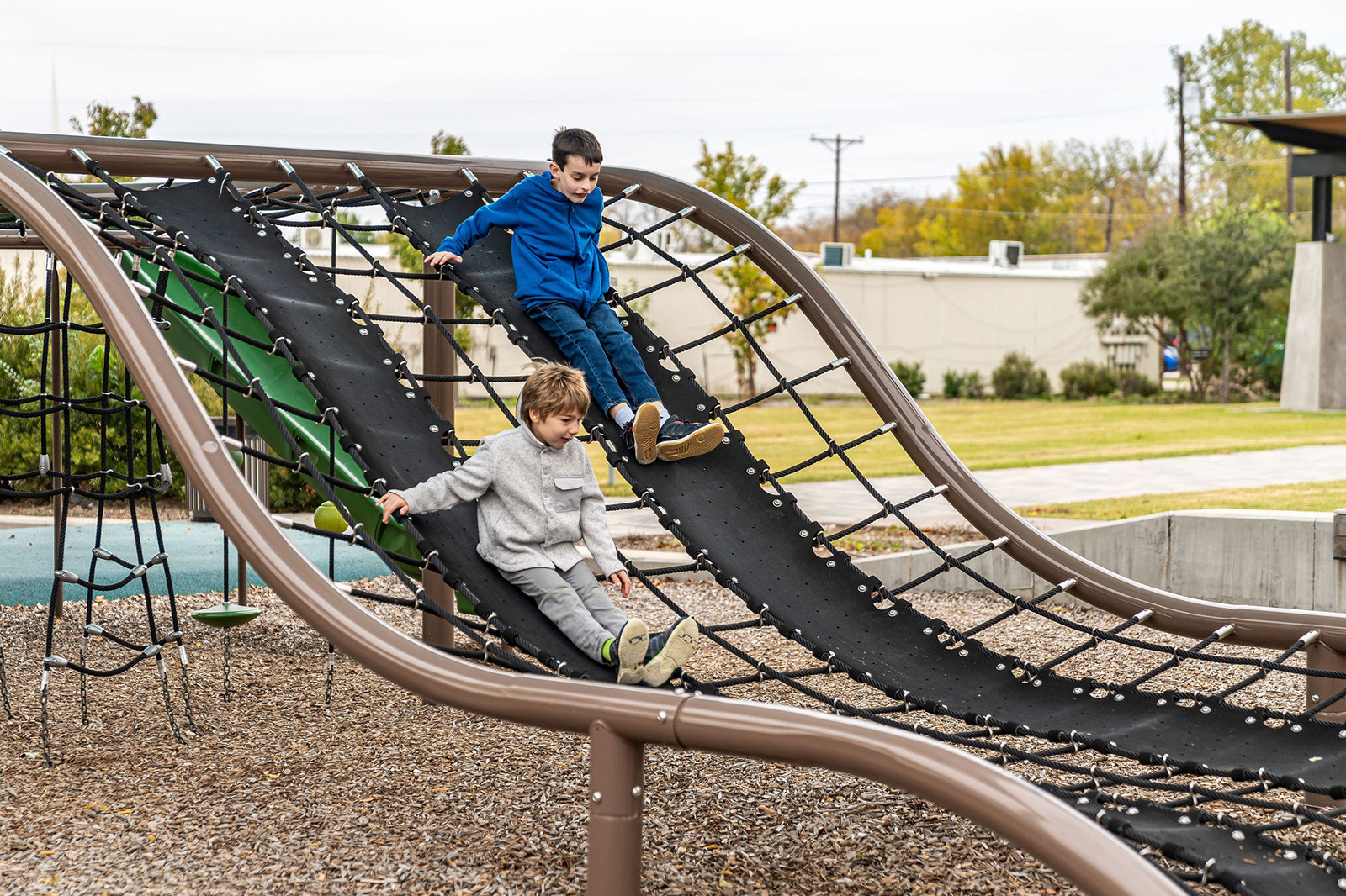 Two boys slide down a black netted climbing structure at an outdoor playground on a cloudy day.