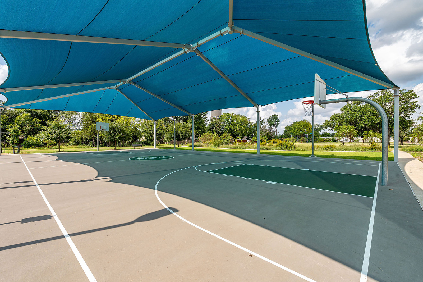 Covered outdoor basketball court with a blue shade canopy, multiple hoops, and clear weather in a park setting.
