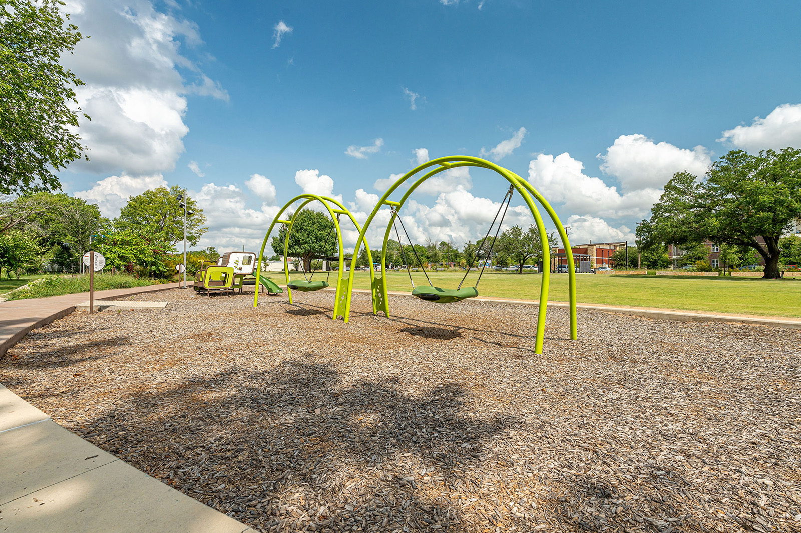 Two green swings are positioned in a playground area with wood chip groundcover, surrounded by grass, trees, and a partly cloudy sky.