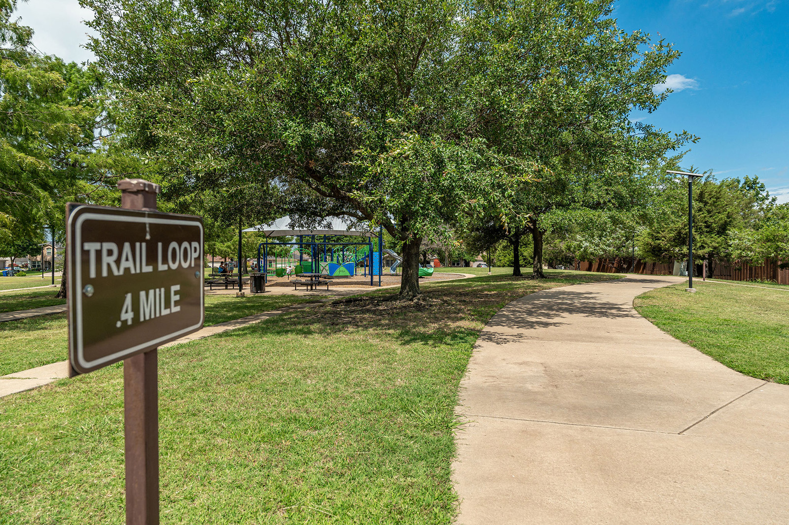 Paved walking trail loops through a park with a sign reading "TRAIL LOOP .4 MILE," trees, grass, and a playground visible in the background.