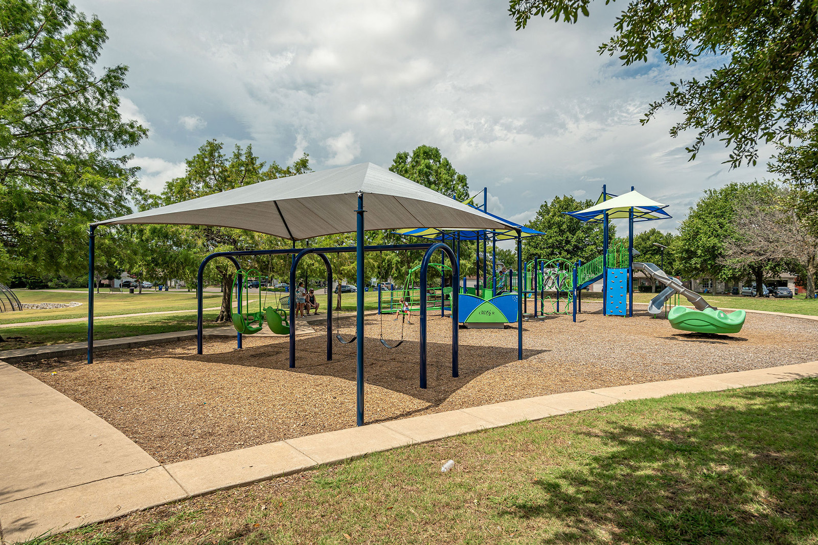 Playground with swings, slides, climbing structures, and a shade canopy, surrounded by trees and grass under a partly cloudy sky.