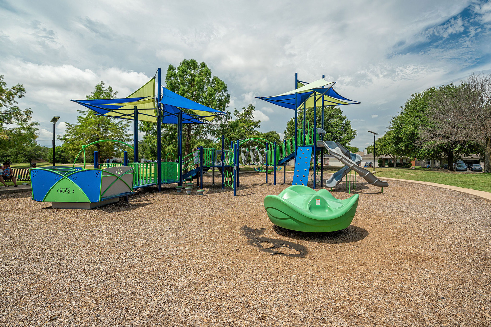 Outdoor playground with blue and green equipment, slides, climbing structures, and wood chip ground cover, surrounded by trees on a cloudy day.
