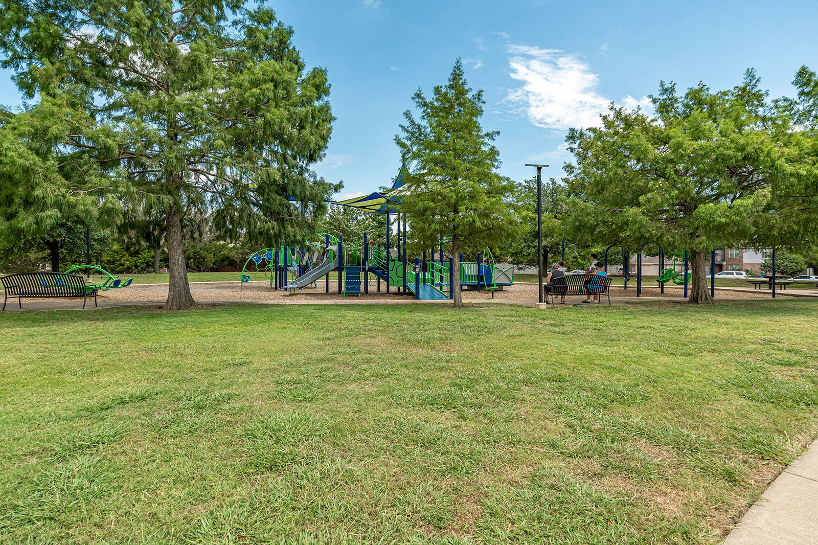 A playground with slides and climbing structures is surrounded by trees, benches, and open grassy areas under a partly cloudy sky.