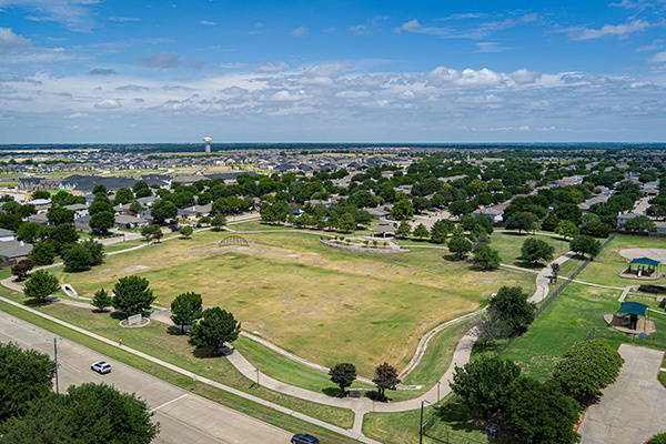 Aerial view of a neighborhood park with open grassy areas, scattered trees, walking paths, and surrounding residential houses under a partly cloudy sky.