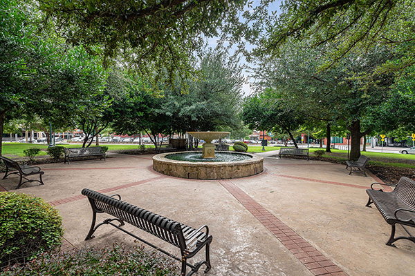 A circular stone fountain sits at the center of a small park surrounded by benches, trees, and a paved walkway.