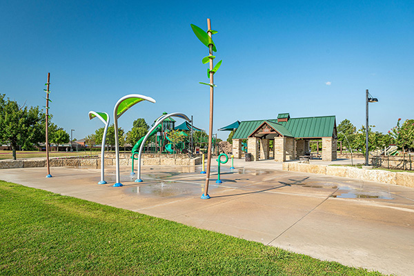 An outdoor splash pad with green and white water features is shown near a grassy area, with a stone pavilion and playground visible in the background under a clear blue sky.