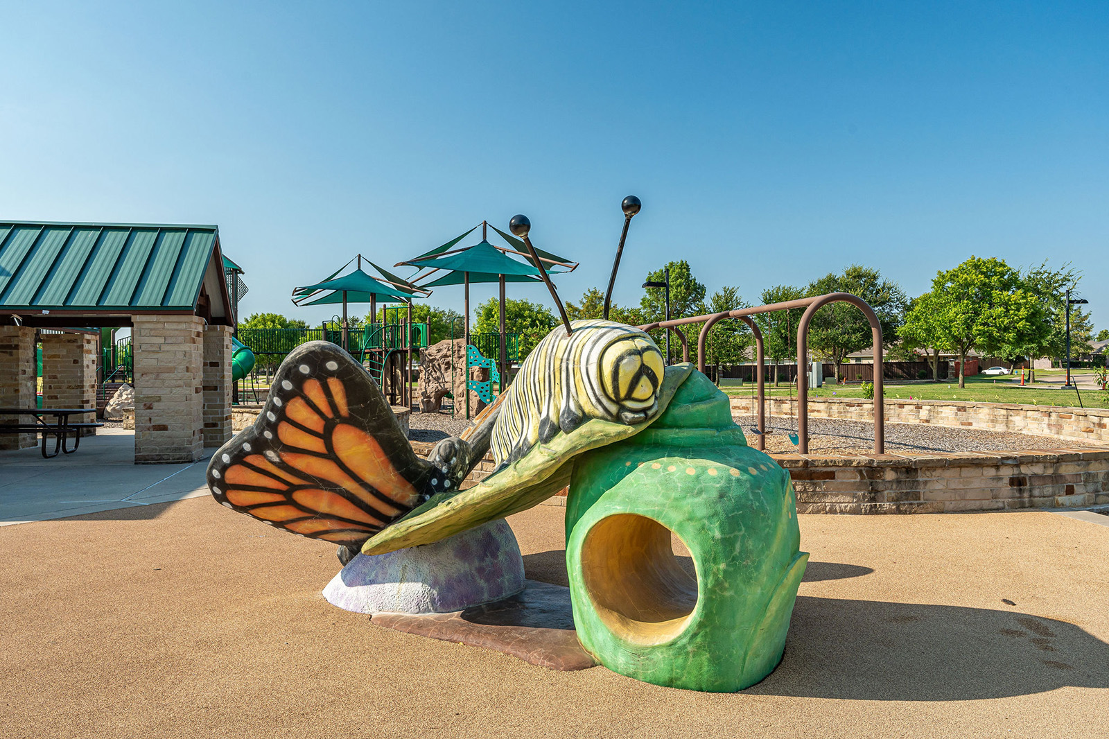 Colorful playground structure shaped like a caterpillar and butterfly, with slides and climbing areas, set on a rubber surface near covered picnic tables and green trees.