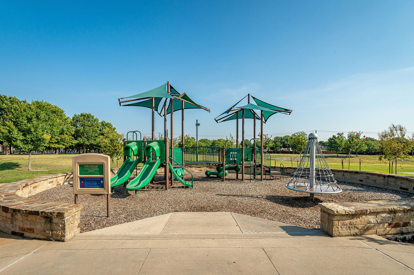 A playground with green slides, climbing structures, and shade canopies is surrounded by trees and grass under a clear blue sky.