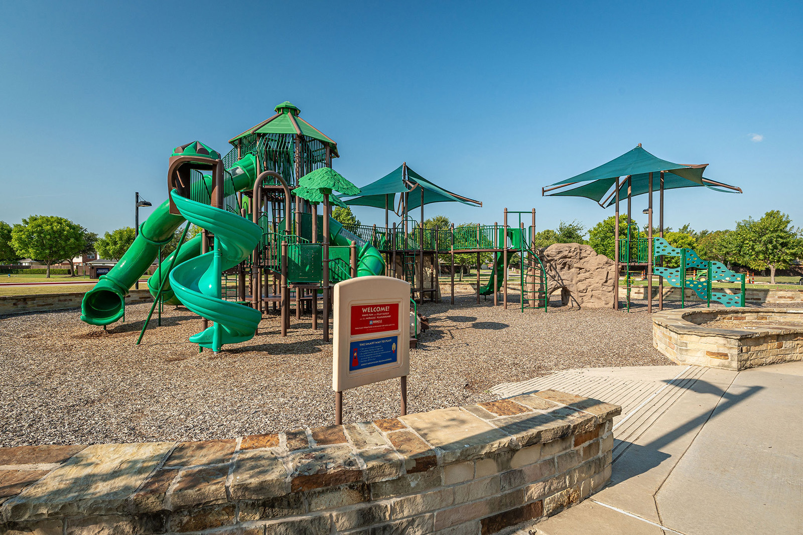 Playground with green slides and climbing structures, shaded areas, a rock climbing wall, and a welcome sign in the foreground, set in a park with trees and blue sky.