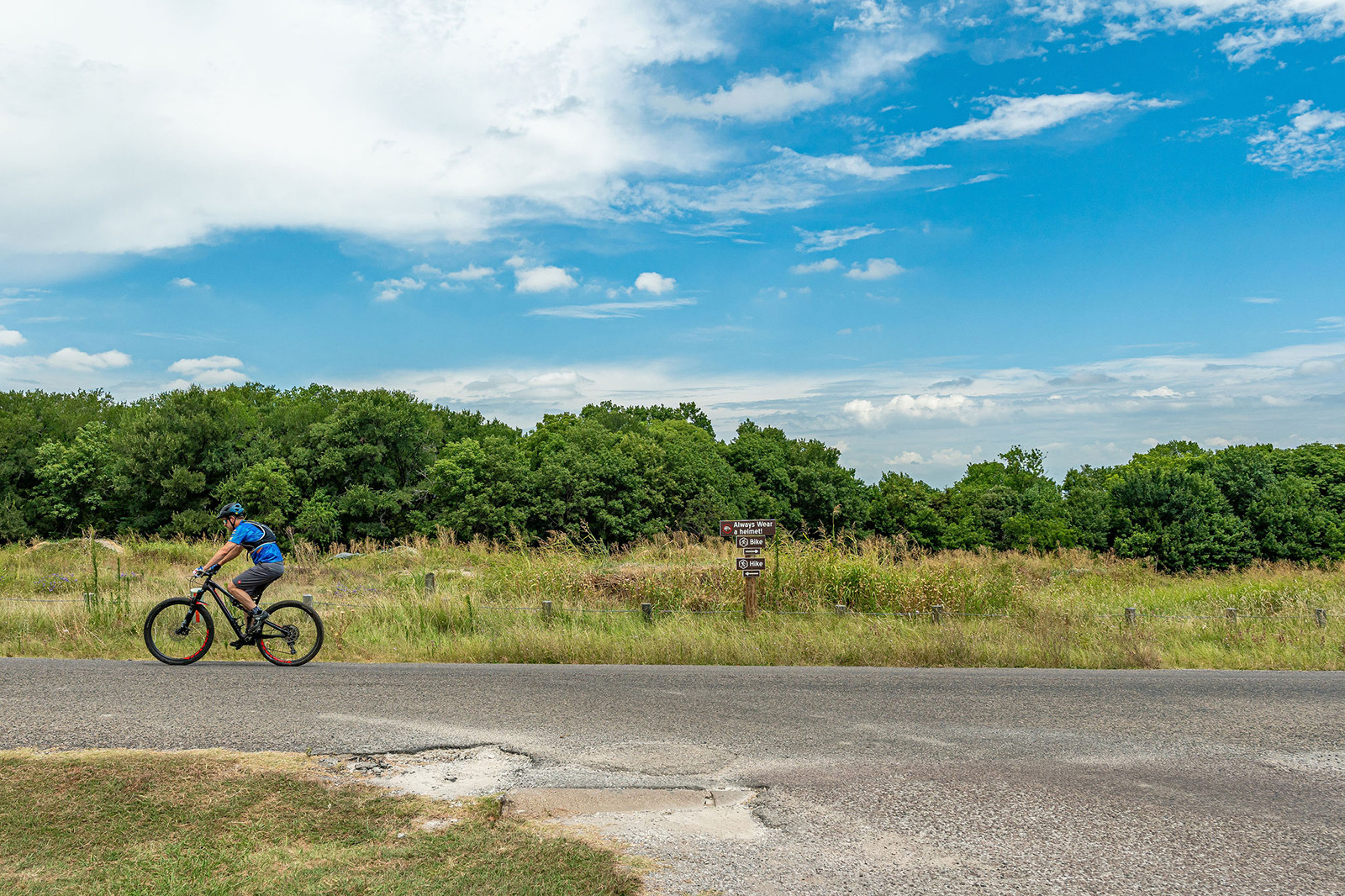 A person wearing a blue shirt and helmet rides a bicycle along a paved road bordered by grass and trees under a partly cloudy sky.