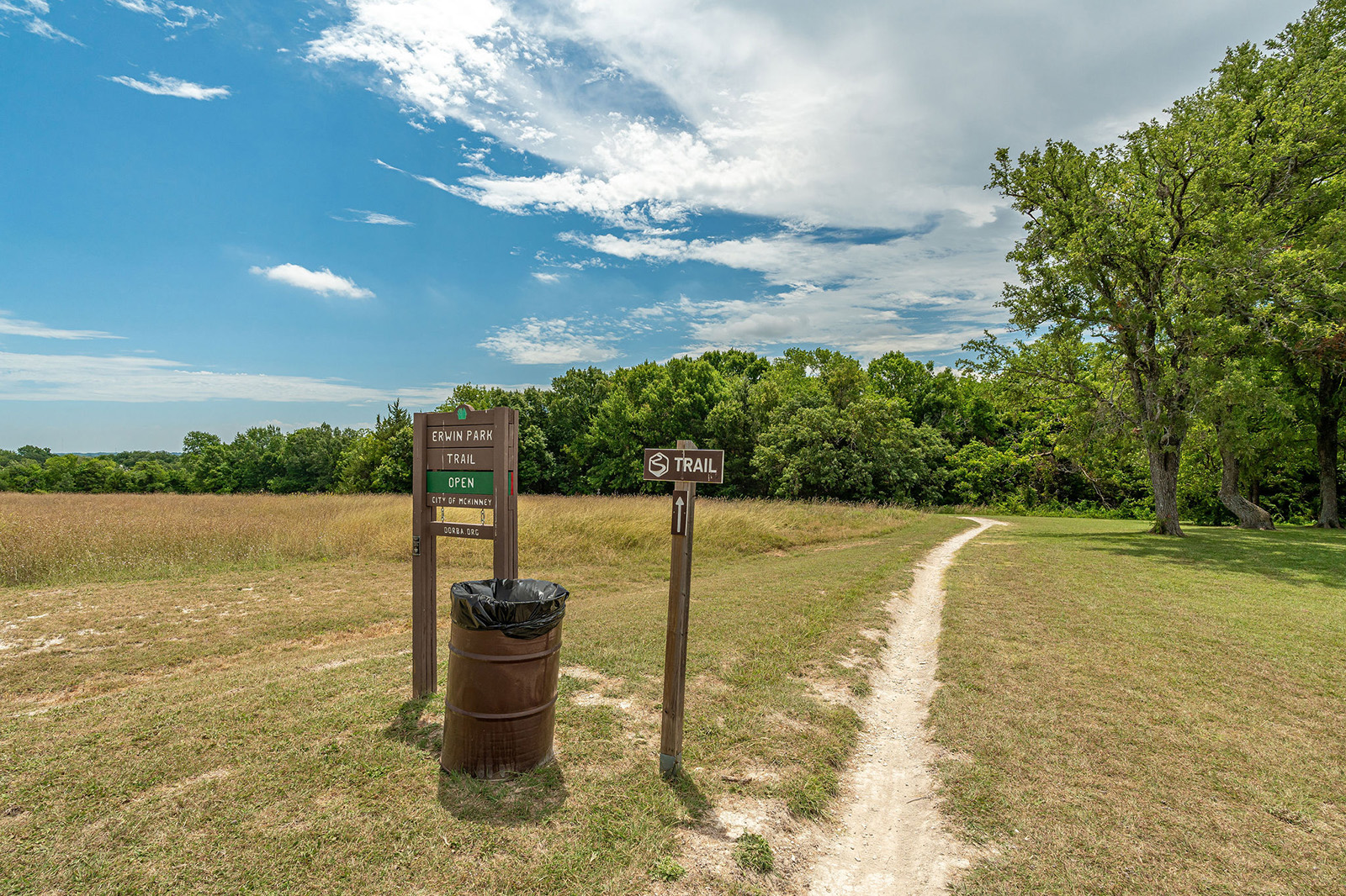 A dirt hiking trail curves through grasslands toward trees, with trail signs and a metal trash can at the entrance under a partly cloudy sky.