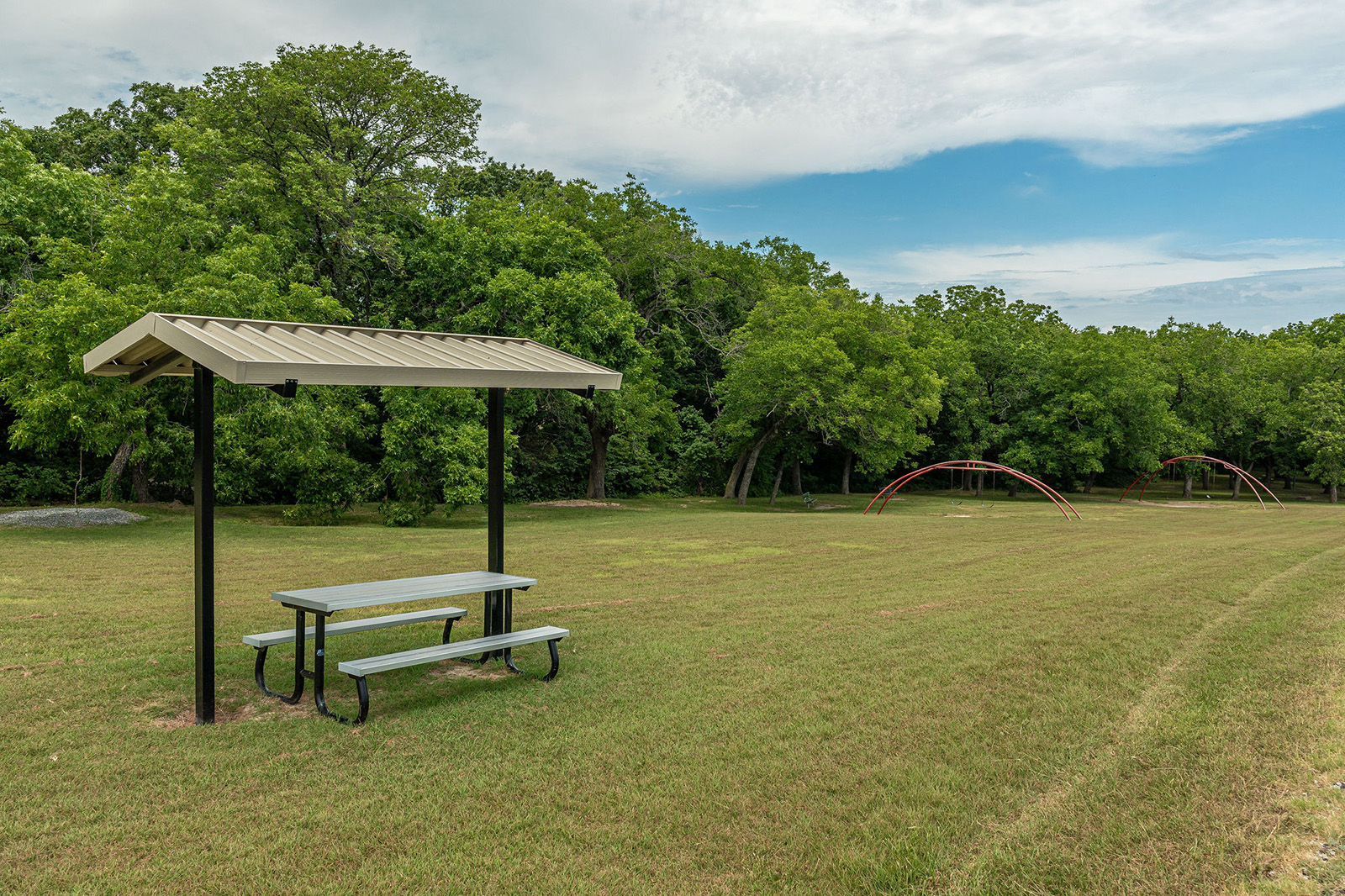 A covered picnic table sits on a grassy field near a tree line, with red playground arches visible in the distance under a partly cloudy sky.