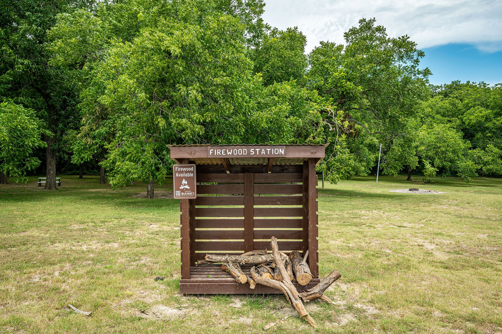 A wooden firewood station with a few logs in front, set in a grassy park area with green trees and a firewood availability sign.