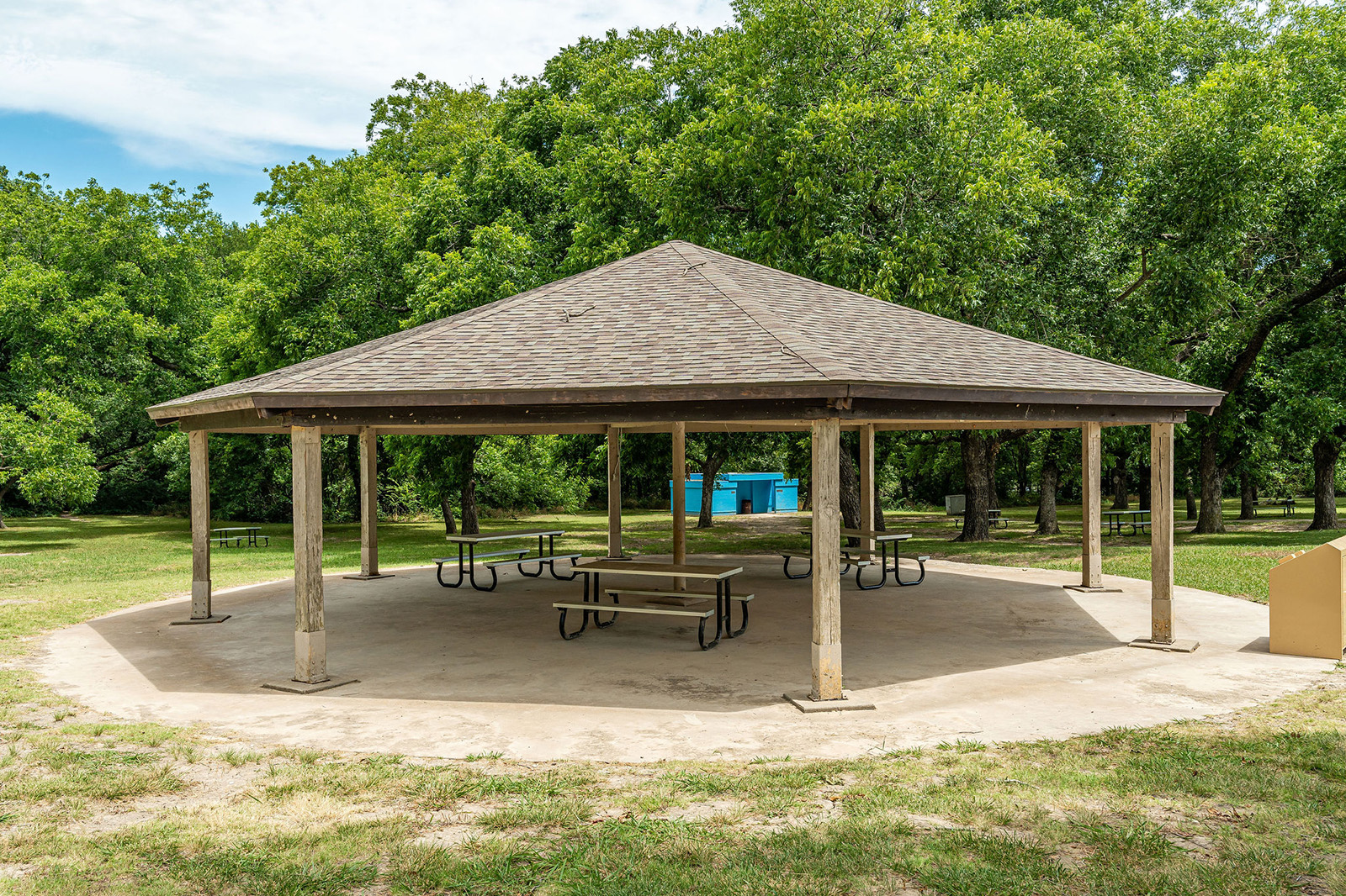 A hexagonal open-air pavilion with a shingled roof covers several picnic tables on a concrete pad in a grassy park area with trees in the background.