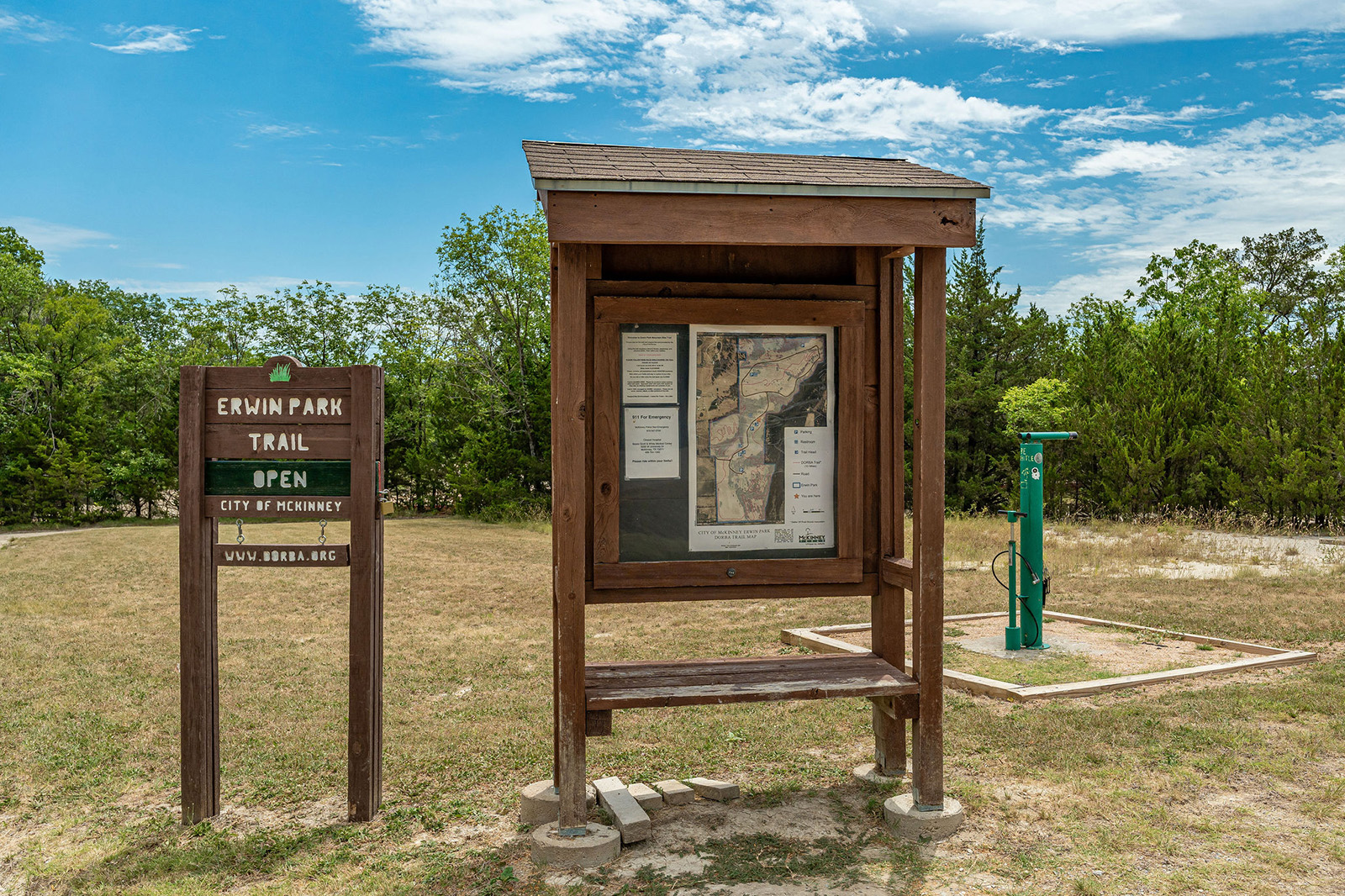 A wooden information board with a map stands next to a green trail sign and water station at Erwin Park Trail under a partly cloudy sky.
