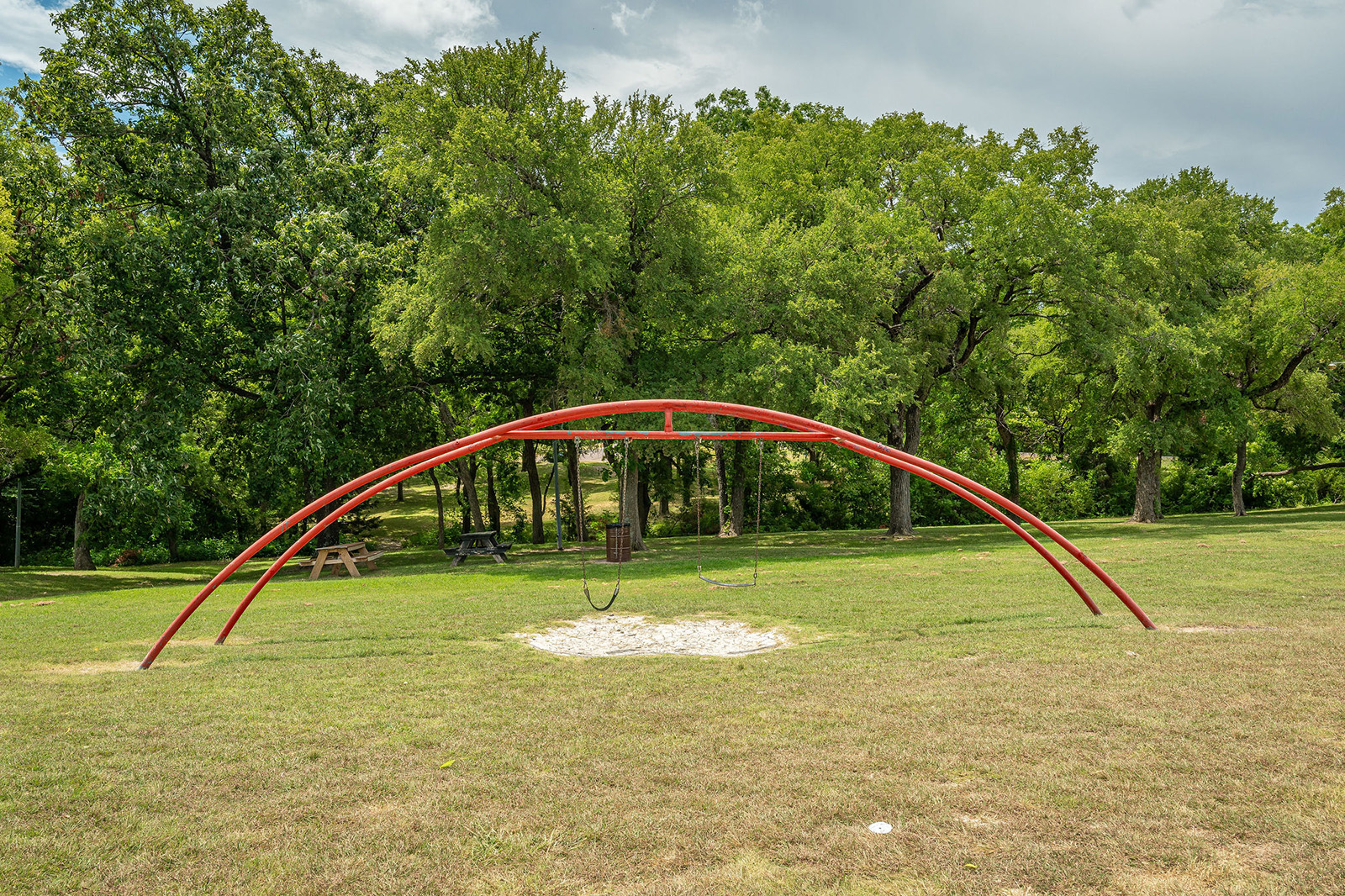 A red metal swing set with two swings stands on a grassy field, with trees and picnic tables in the background.