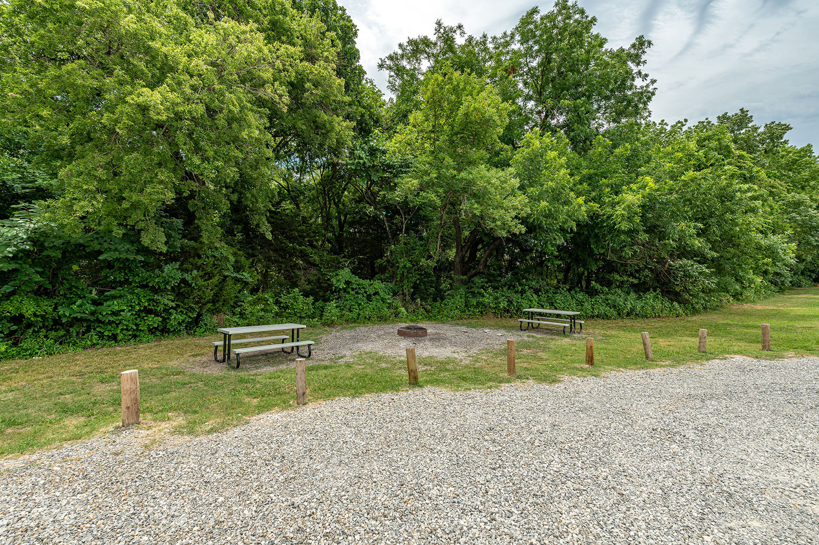 Two picnic tables near a fire pit on a gravel area, bordered by wooden posts, with dense green trees in the background.