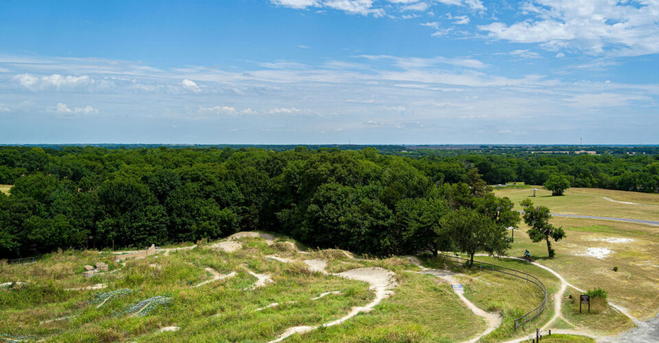 A winding dirt trail cuts through grassy terrain with scattered trees, bordered by a dense forest under a partly cloudy blue sky.