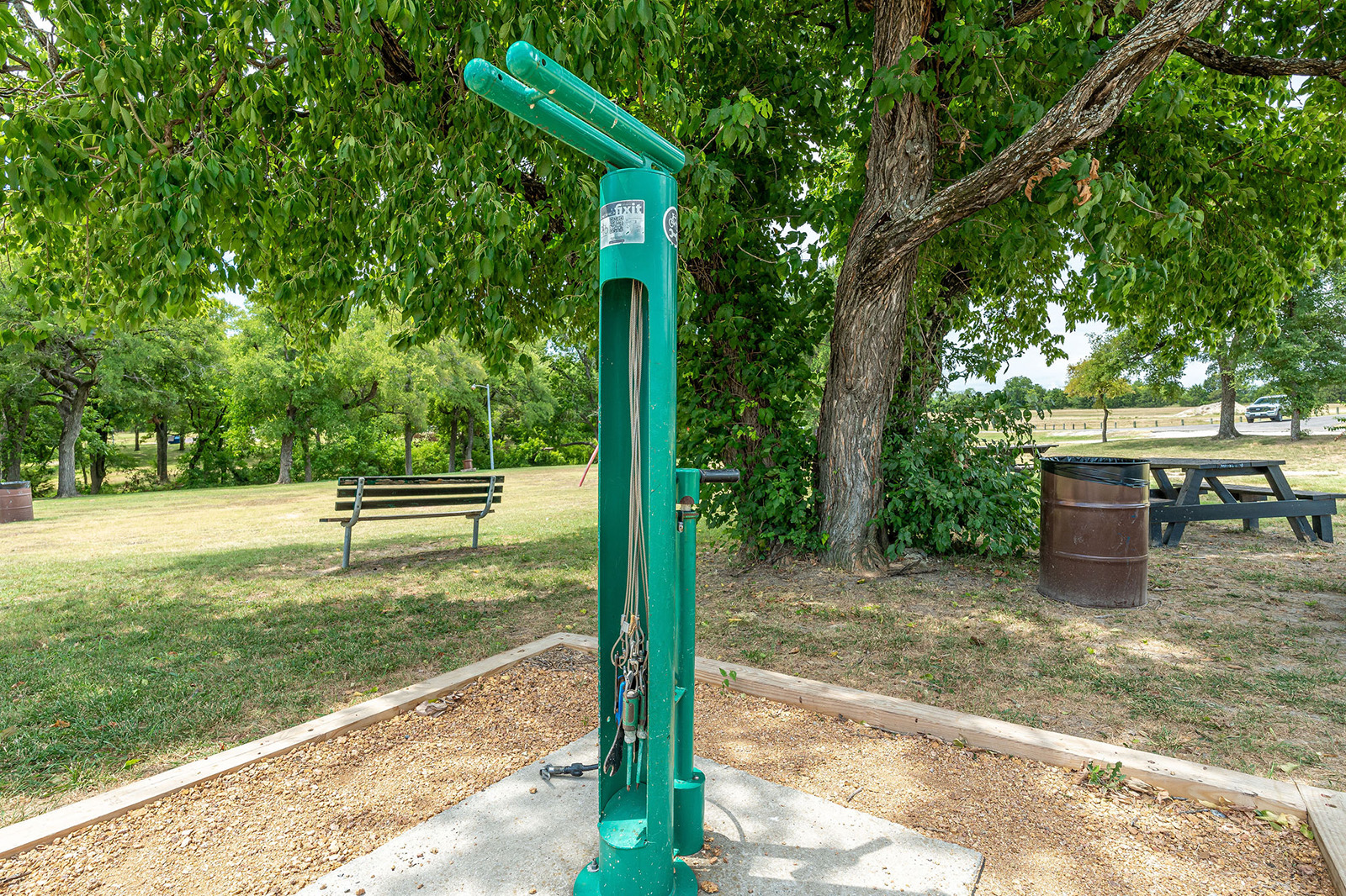 Green outdoor repair station with tools attached, located near a bench and picnic table in a park setting with trees and grass.