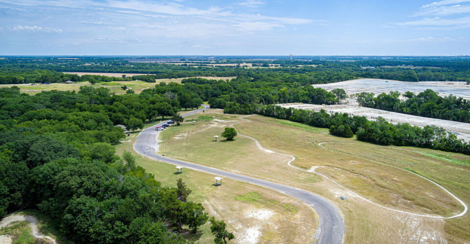 A winding paved road curves through a grassy, open park area with scattered trees, surrounded by dense green forest under a partly cloudy sky.