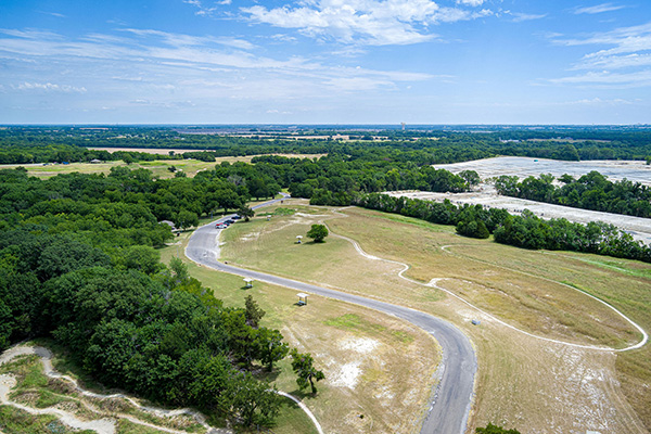 A winding paved road runs through open grassy fields and scattered trees under a partly cloudy sky, viewed from above.