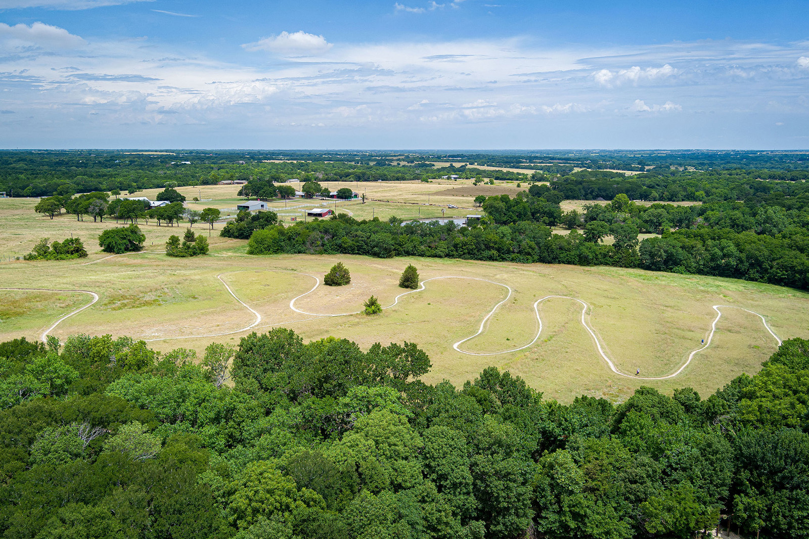 Aerial view of a winding dirt trail through an open grassy field bordered by trees, with farms and forest in the background under a partly cloudy sky.