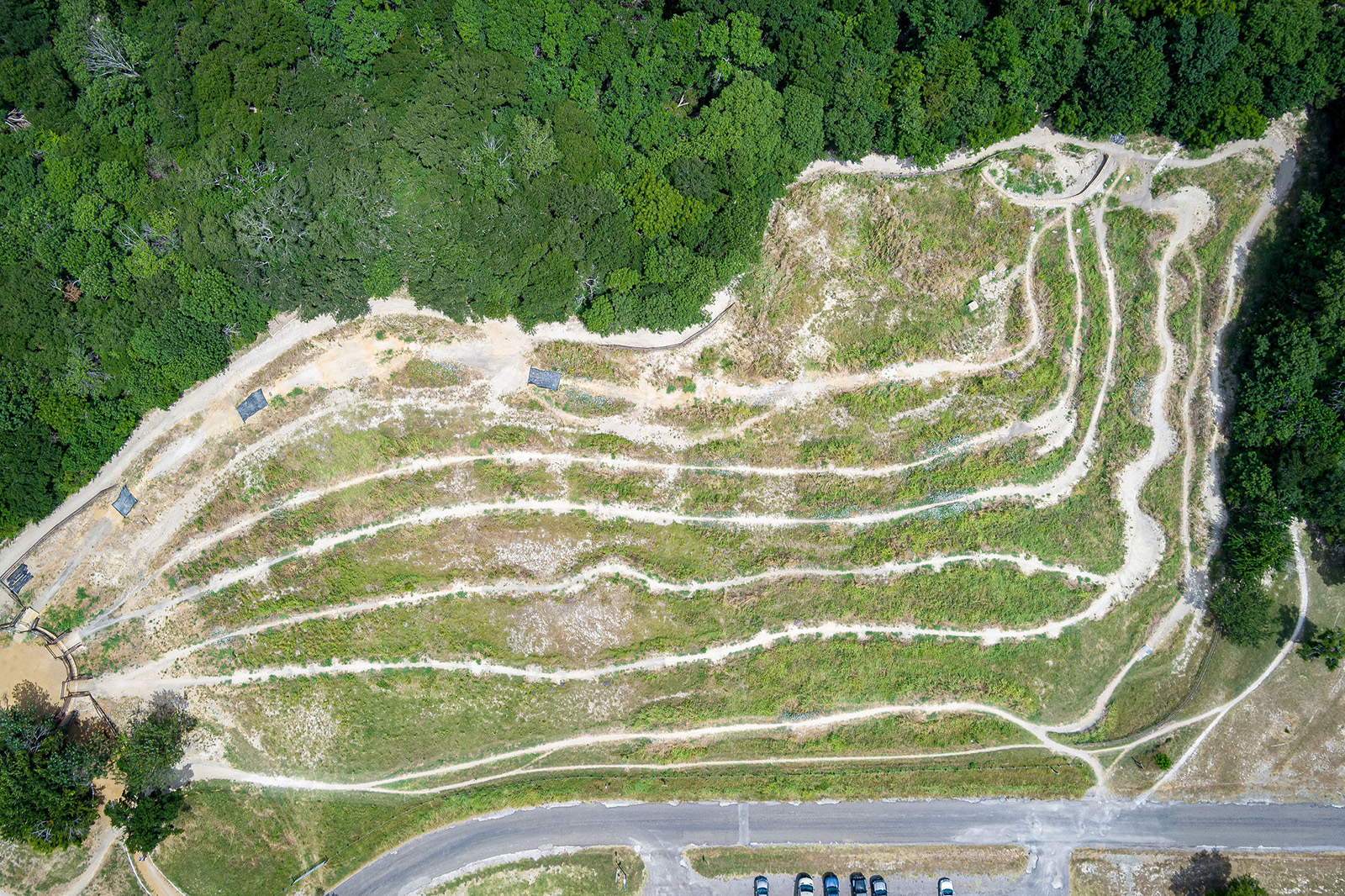 Aerial view of a terraced hillside with winding dirt paths near a forested area, adjacent to a paved parking lot with parked cars.
