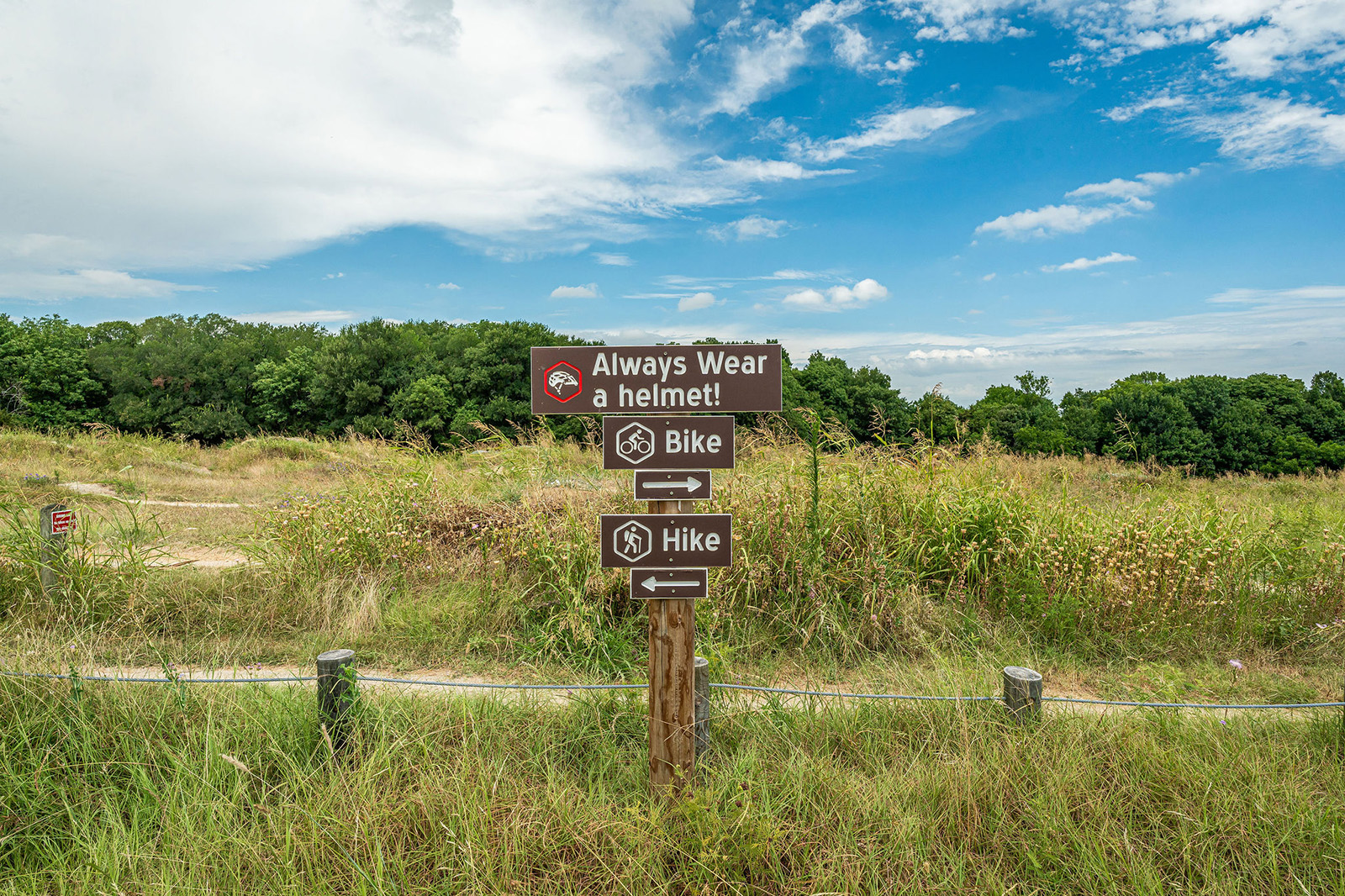 Wooden trail sign in grassy field shows directions for biking left and hiking right, with a reminder to always wear a helmet. Trees and blue sky in the background.