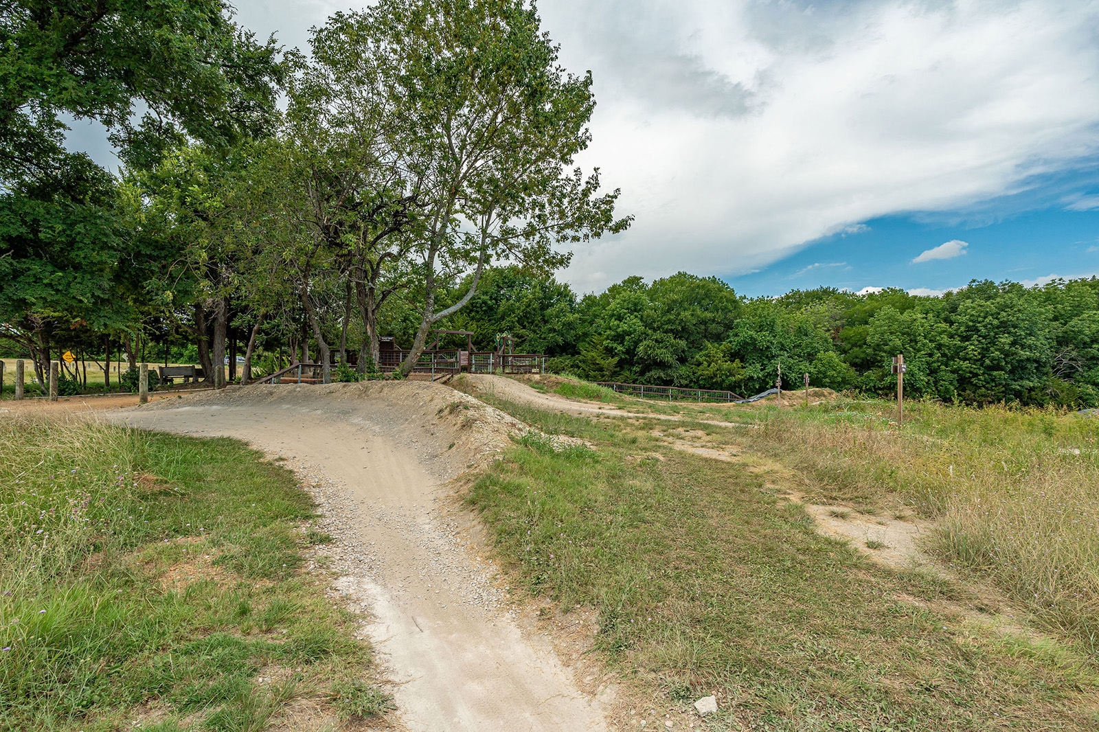Dirt path curves through grassy area with scattered trees under a partly cloudy sky. Wooden fence and small structure are visible in the background.
