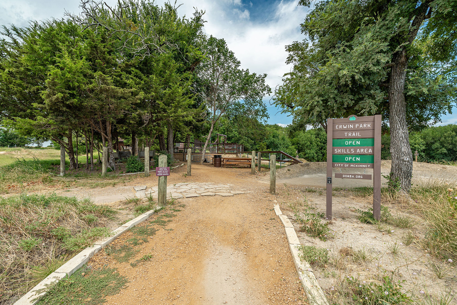 A dirt pathway leads to a wooded area with benches and signposts indicating Erwin Park Trail and Skills Area are open. Trees and cloudy sky are visible in the background.