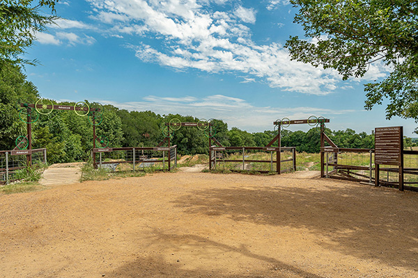 A dirt path leads to several fenced gates with metal signs, surrounded by trees and grass under a partly cloudy sky.