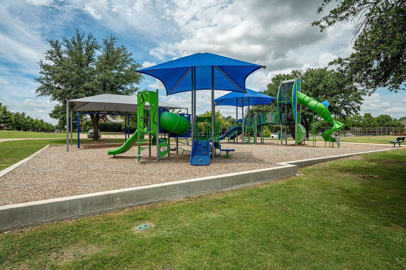 A playground with green and blue climbing structures, slides, and large blue shade canopies, surrounded by grass and trees under a partly cloudy sky.
