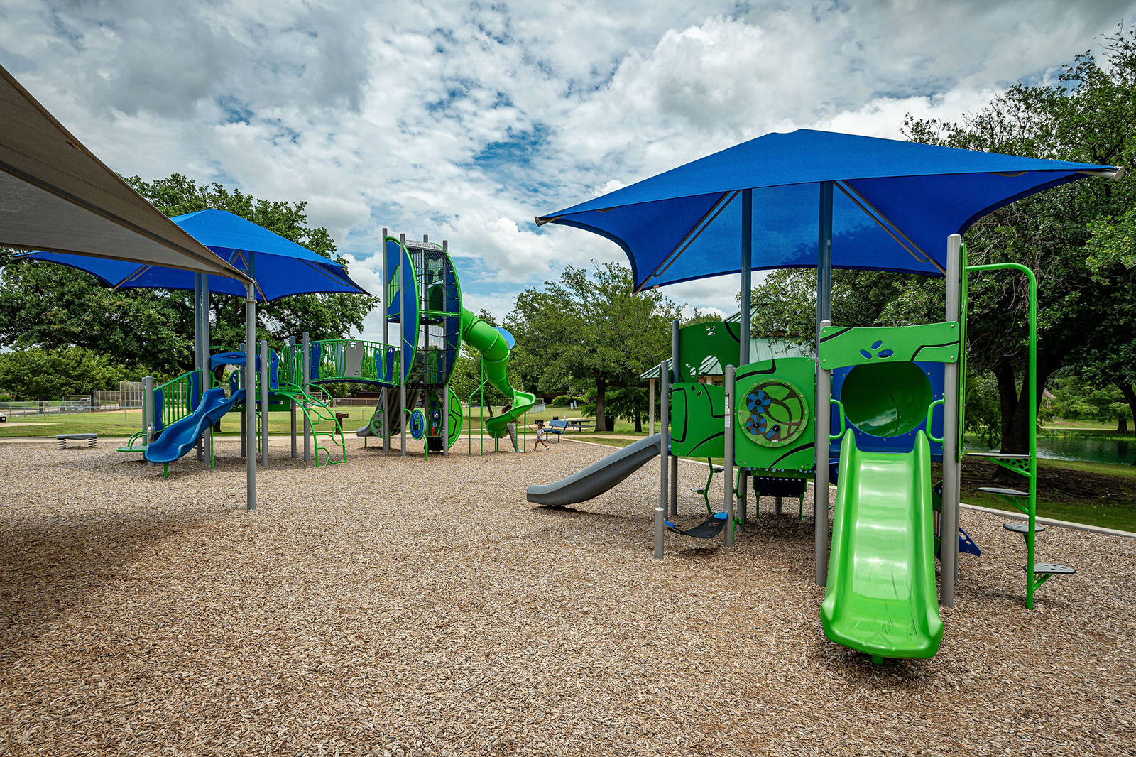 Outdoor playground with green and blue slides, climbing structures, and shade canopies on a mulch surface, surrounded by trees under a cloudy sky.