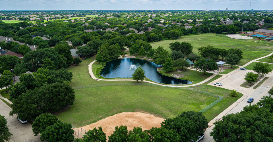 Aerial view of a suburban park with a small pond and fountain, open grassy areas, trees, a baseball field, and surrounding houses under a partly cloudy sky.